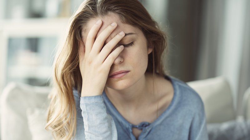 A young woman wearing a long-sleeved pajama top and holding her head with her eyes closed because of exhaustion